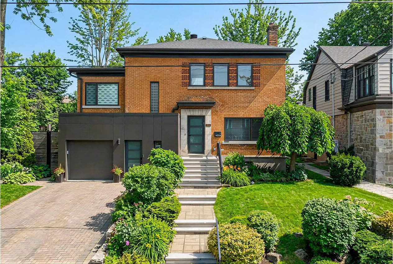 Two-story brick house with a gray garage and front door. A stone walkway leads up to the house, surrounded by green bushes and grass.
