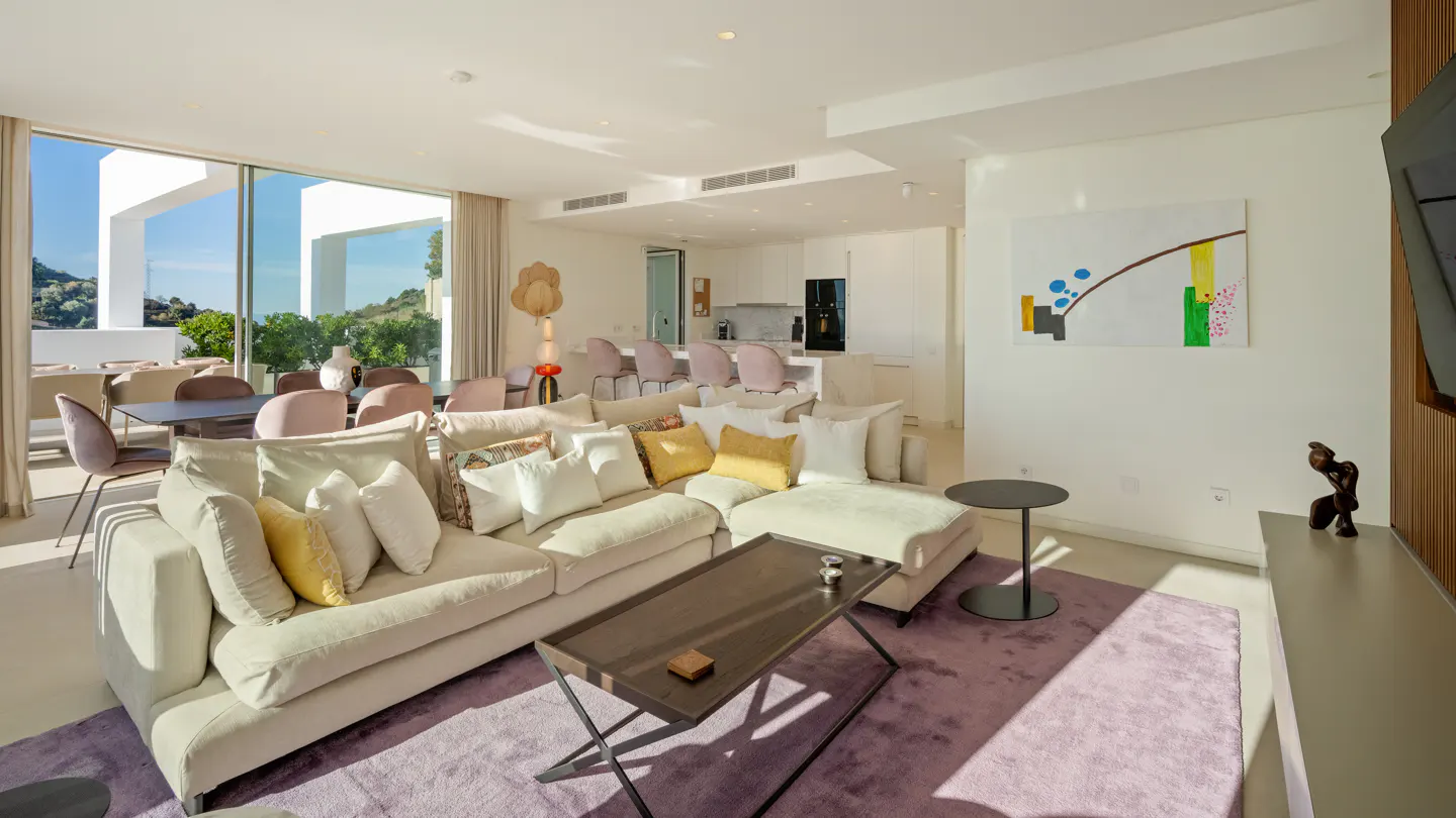 Bright, modern living room with a large beige sofa, yellow pillows, and a dark coffee table on a purple rug. Open floor plan with dining and kitchen areas.