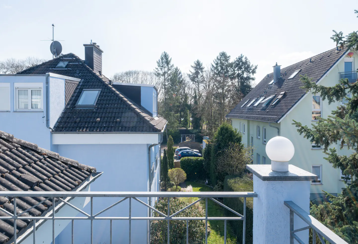 View from a balcony overlooking a residential area with houses, trees, and parked cars on a sunny day.
