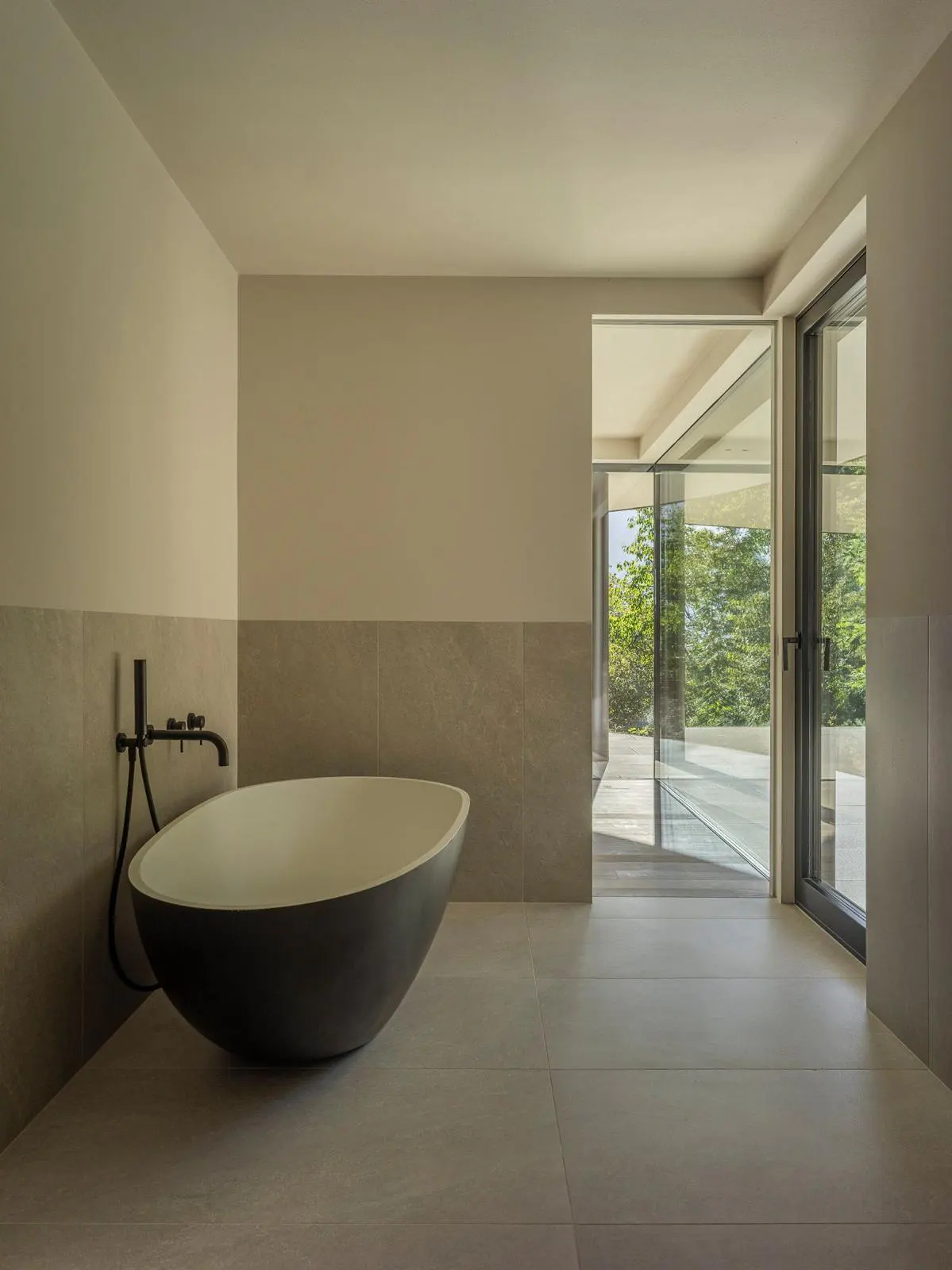 Modern bathroom with a black, oval tub and black faucet. Large glass doors open to a view of green trees. Neutral color scheme.