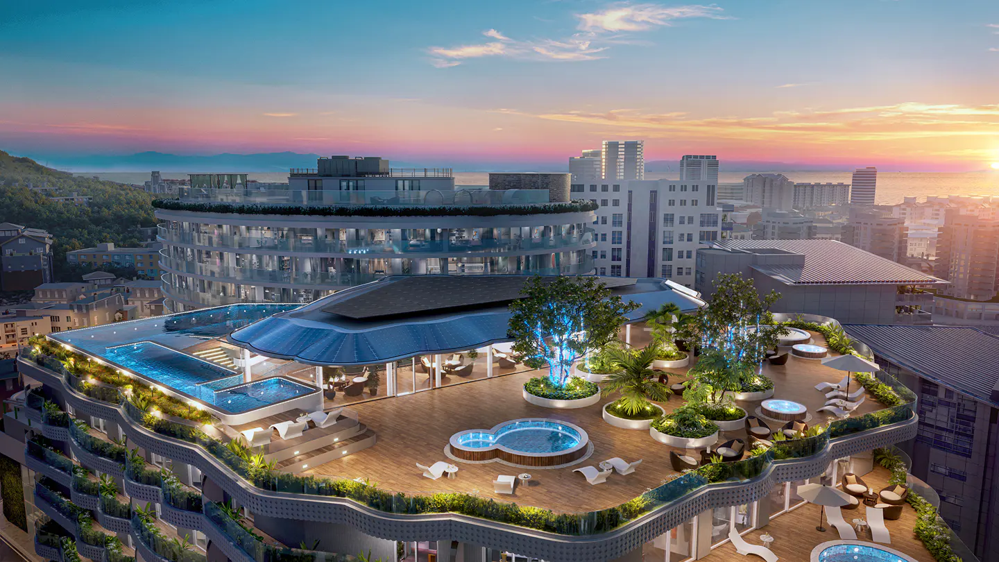 Aerial view of a luxury rooftop terrace with pools, lounge chairs, and greenery at sunset. City buildings and the ocean are in the background.