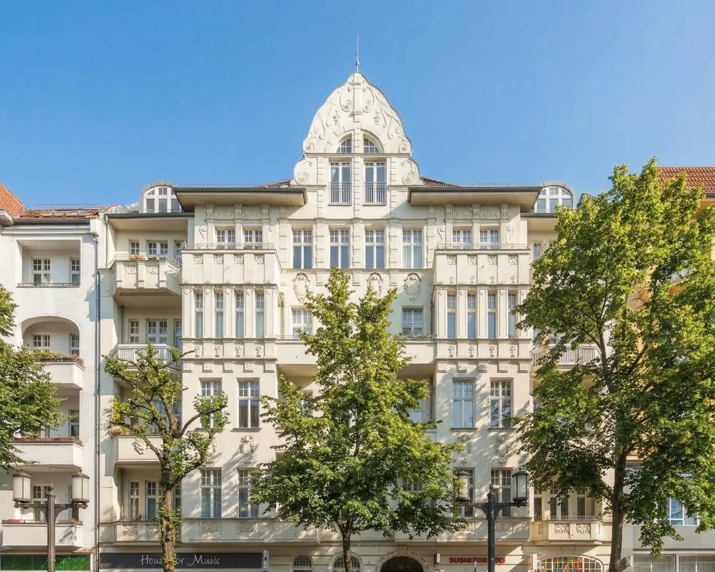 Facade of a light-colored, ornate apartment building with balconies and trees in front on a sunny day.