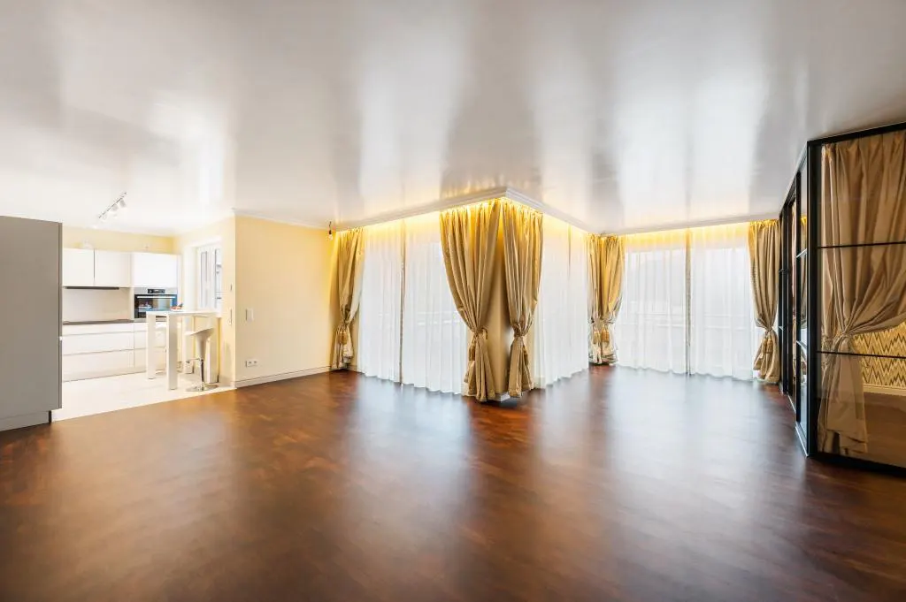 Bright, open living space with dark wood floors, cream walls, and gold curtains. Kitchen visible in the background.