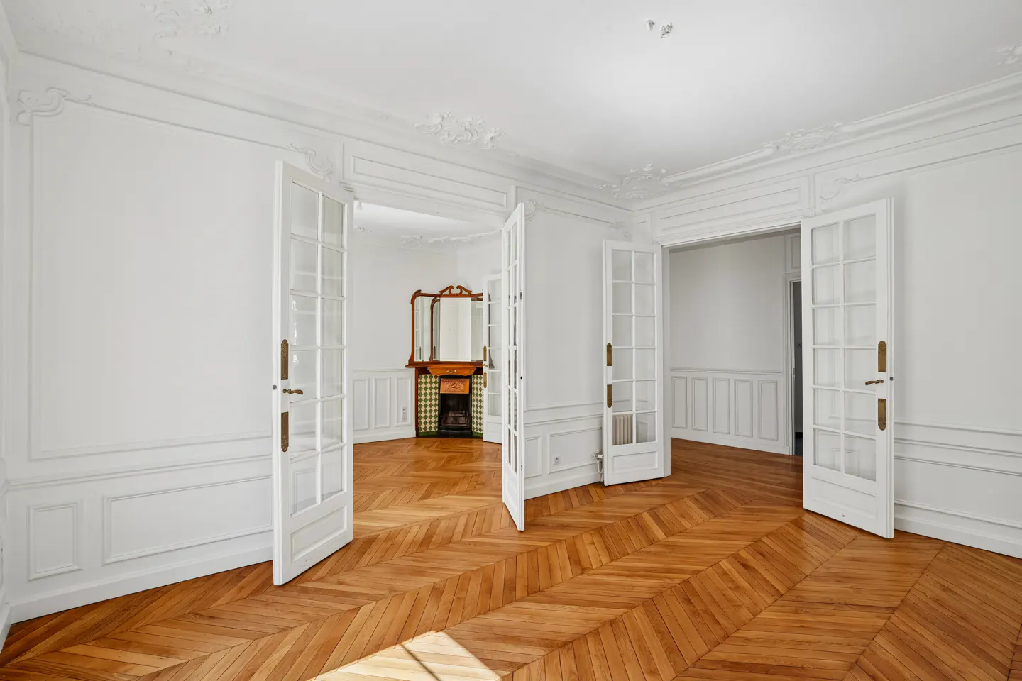 Bright room with white paneled walls, herringbone wood floors, and open glass-paned doors leading to an adjacent room.