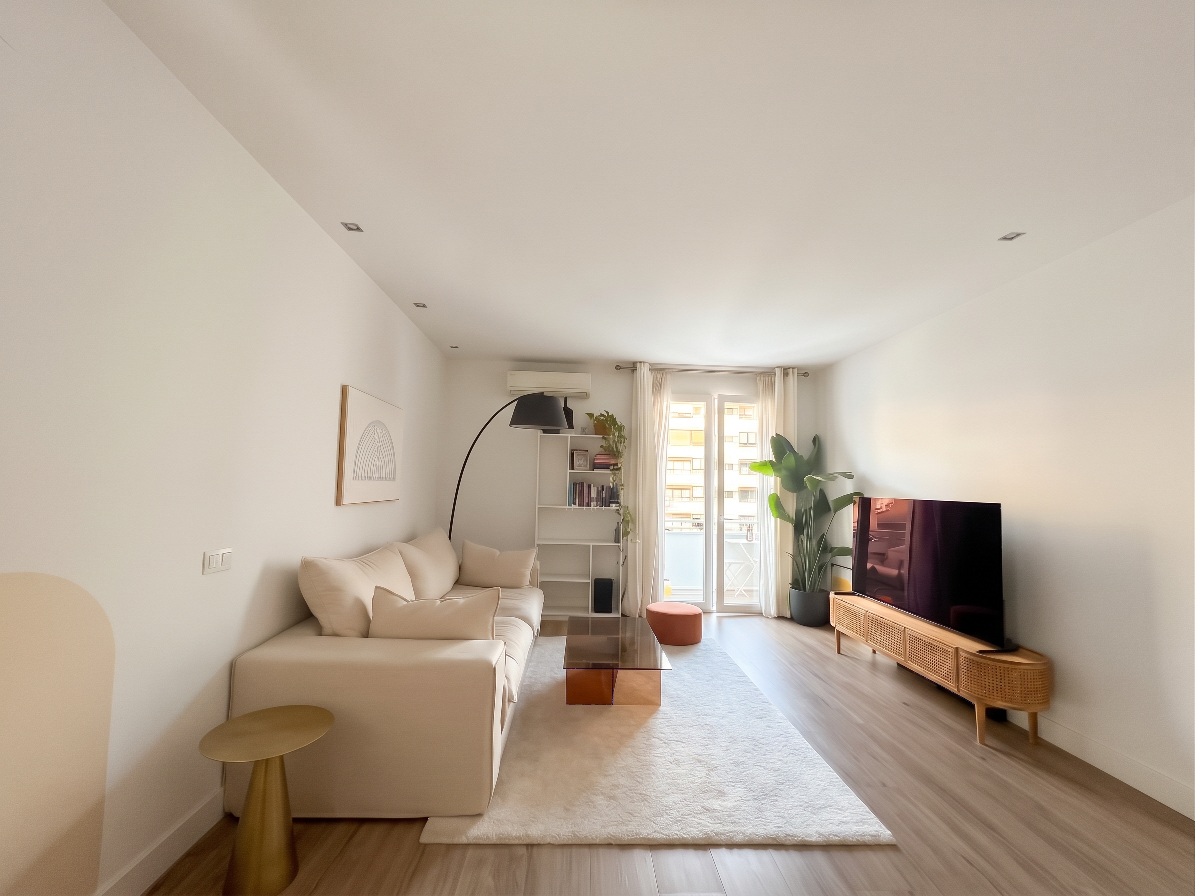 Bright living room with a beige sofa, glass coffee table, TV on a rattan console, and a large plant by a balcony door. [AI-generated text]