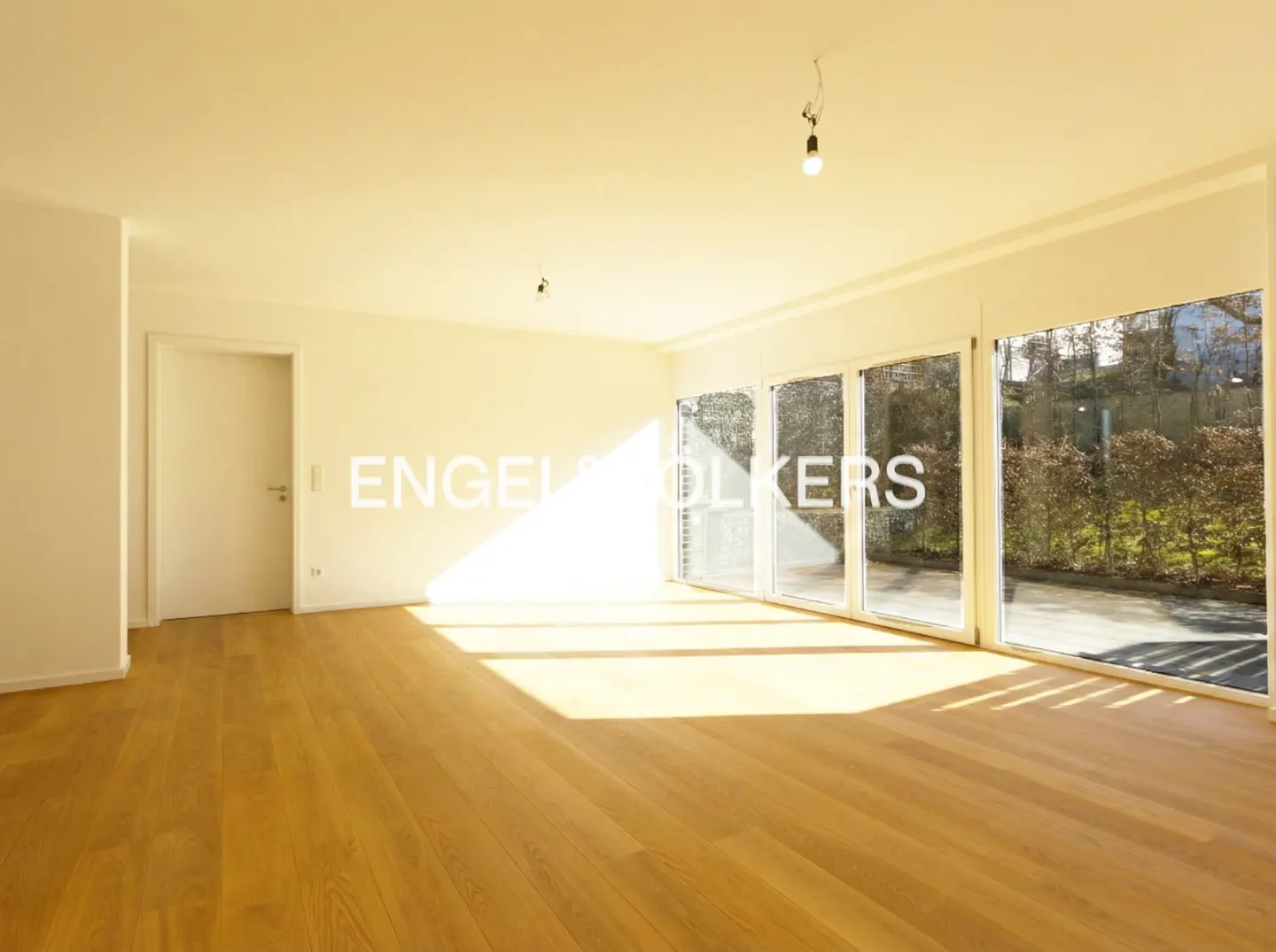 Bright, empty room with light wood floors and white walls. Large windows offer a view of greenery outside.