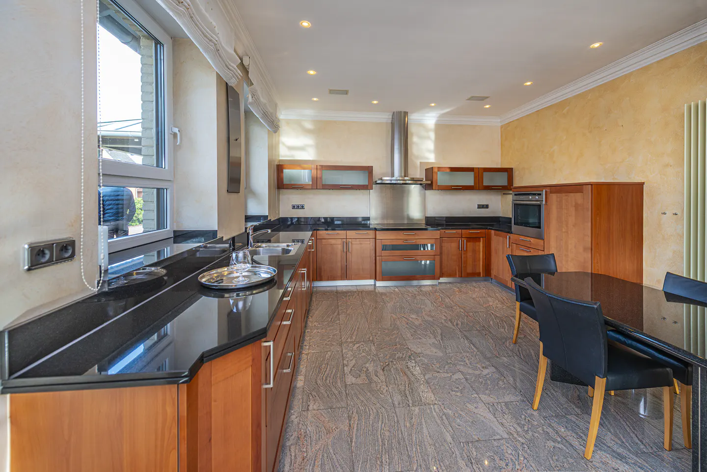 A modern kitchen with wood cabinets, black countertops, and stainless steel appliances. A black table and chairs are in the foreground.