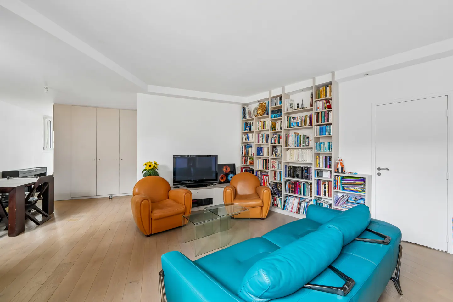 Bright living room with a blue sofa, two orange armchairs, and a large bookshelf filled with books.