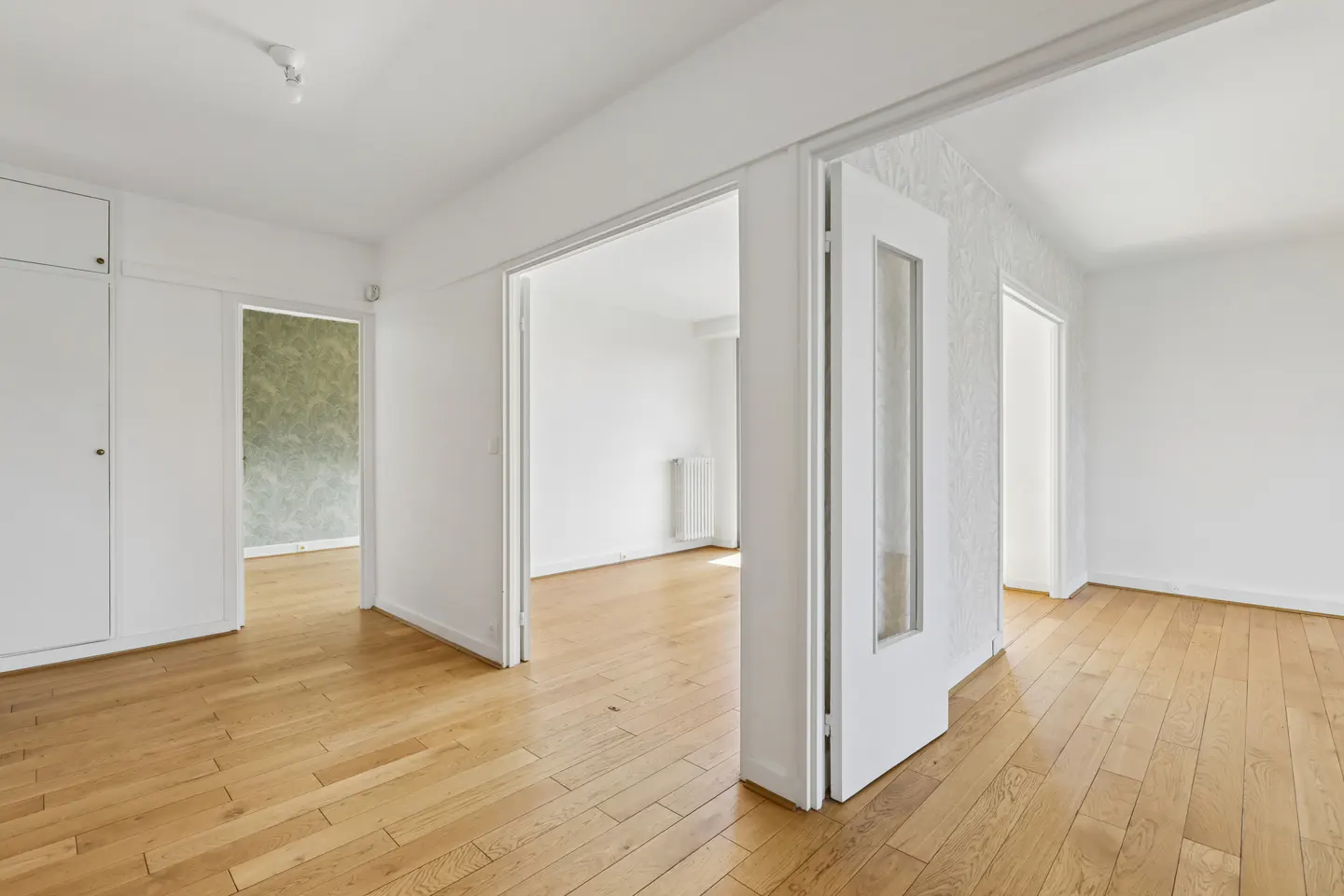 Bright, empty room with hardwood floors and white walls. Open doorways lead to other rooms, one with patterned wallpaper.