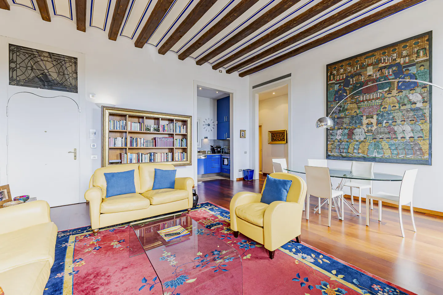 Bright living room with yellow sofas, blue pillows, and a red rug. A glass table sits in front of a bookshelf and a dining area with a large painting.