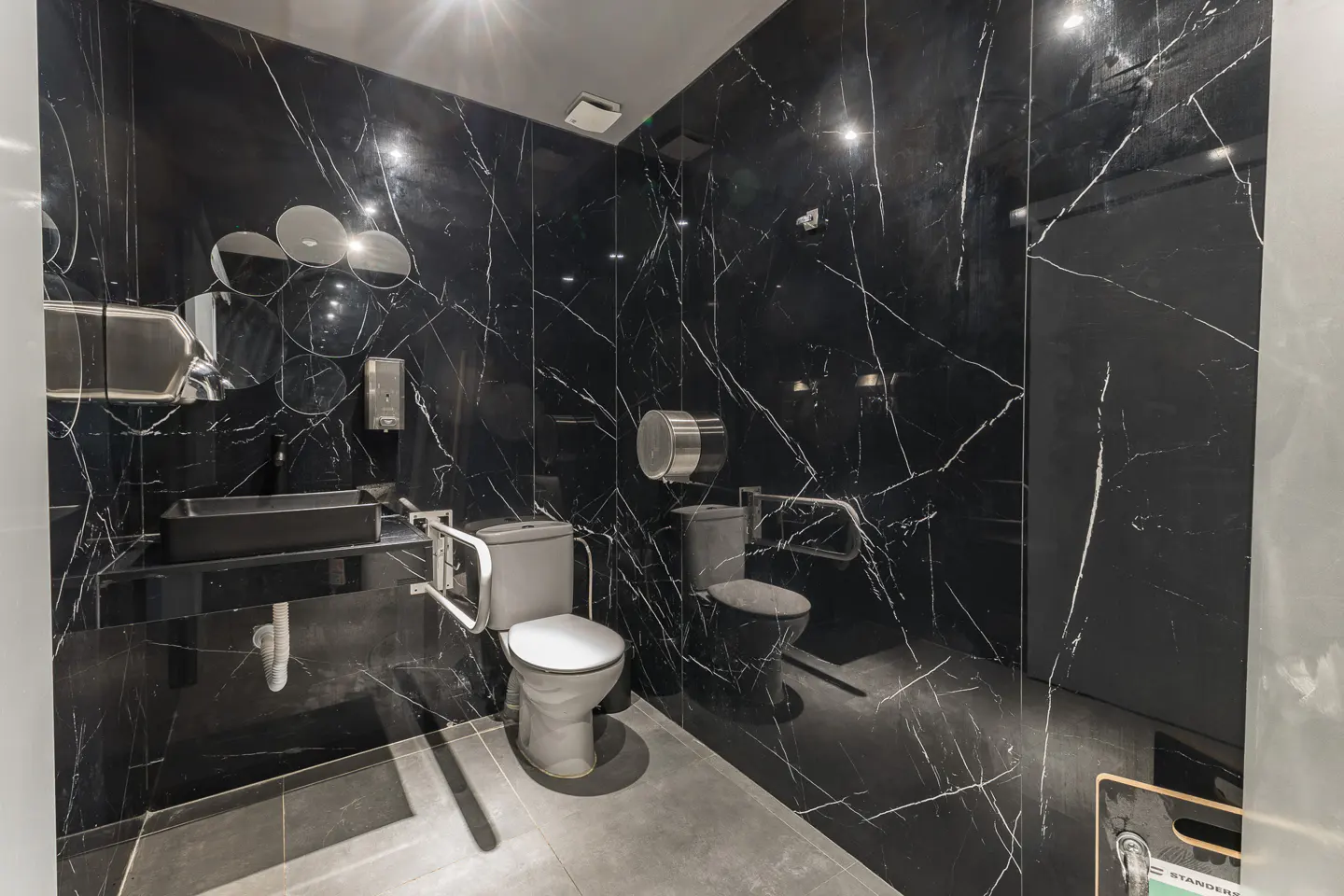 Modern bathroom with black marble walls, gray floor, toilet with grab bars, and a black sink.