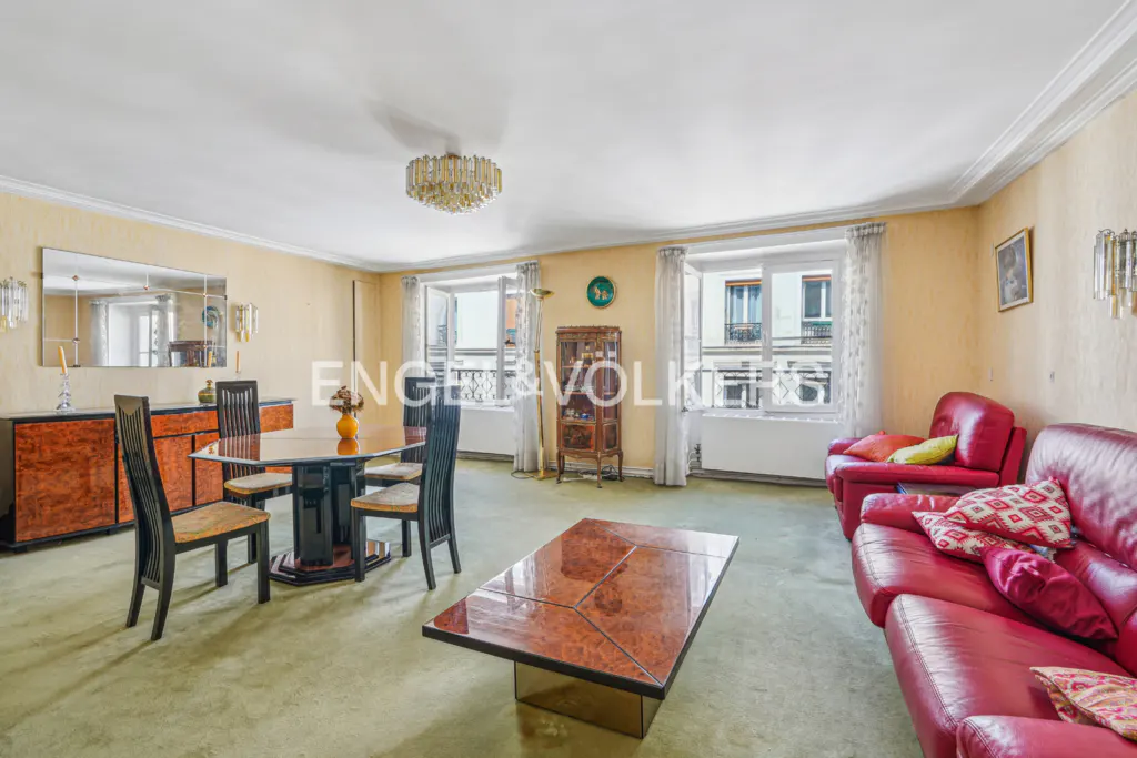 A living room with a dining table, red sofas, and a coffee table. Windows overlook a city.