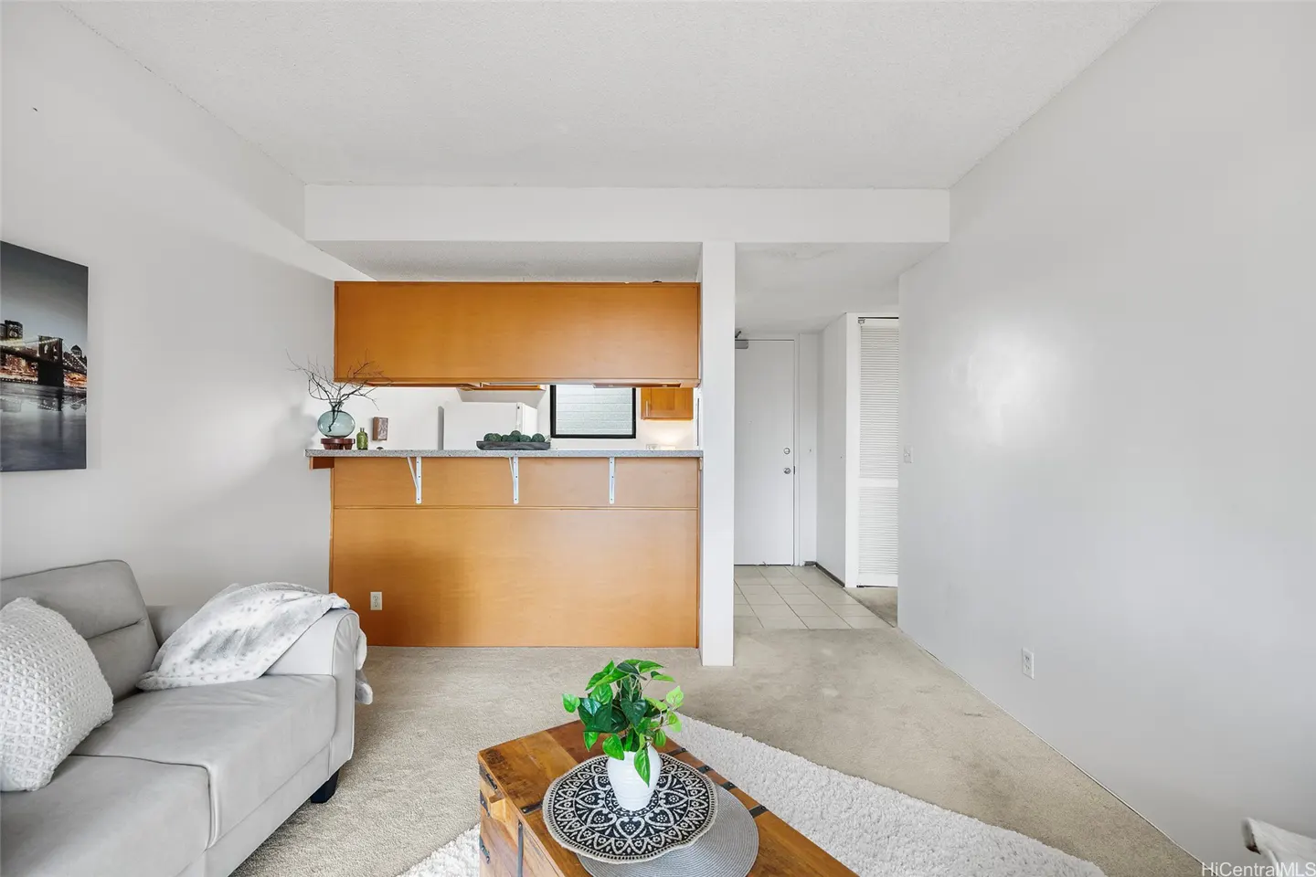 Living room with a gray sofa, wood coffee table, and a view into the kitchen with wood cabinets. The walls are white.