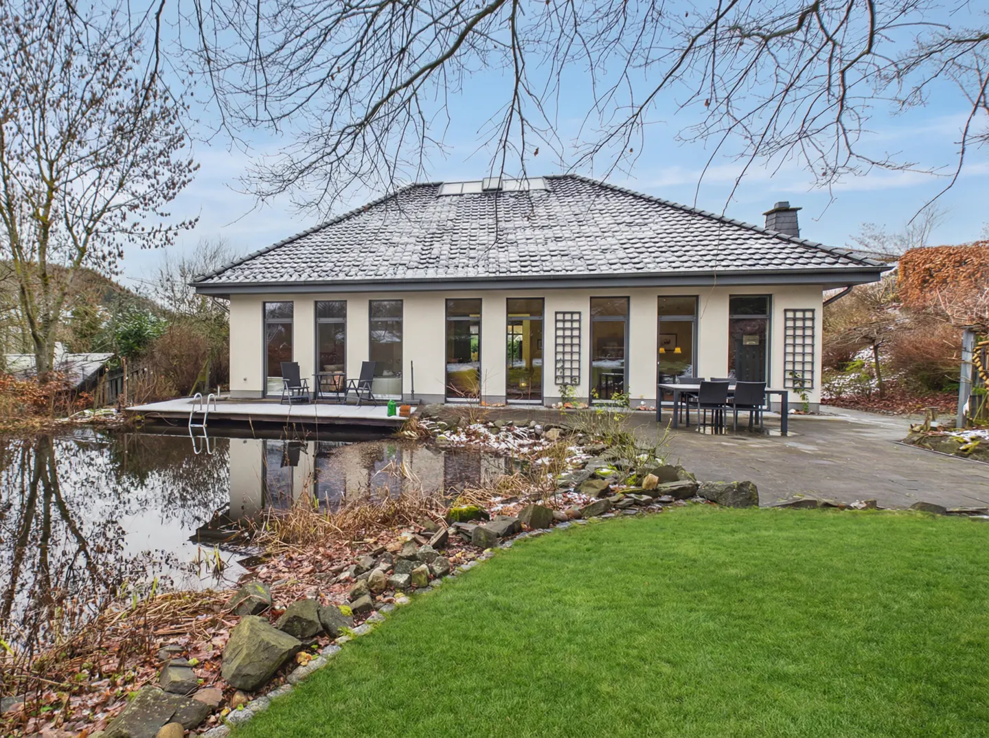 A single-story house with a gray roof overlooks a pond. A wooden deck with chairs extends over the water. Green lawn and trees surround the property.