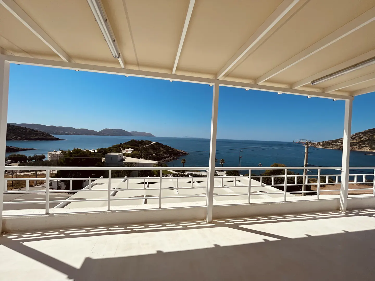 View from a white balcony with a beige awning, overlooking a blue sea and islands under a clear blue sky.