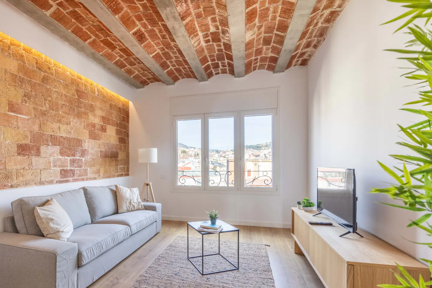 Living room with brick wall, vaulted brick ceiling, gray sofa, wood floors, and a window with a city view.