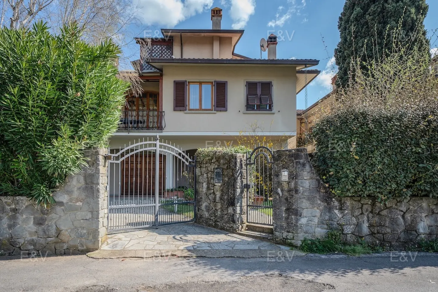 A two-story beige house with brown shutters, a balcony, and a stone wall with metal gates.