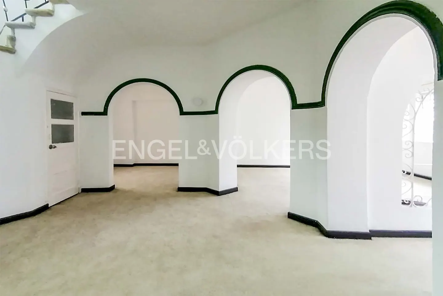 Bright room with white walls, arched doorways trimmed in green, and a beige floor. A white door and stairs are visible.