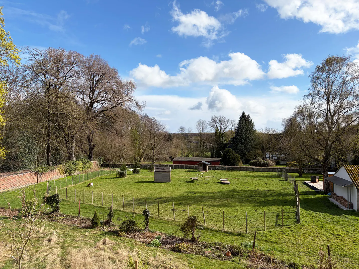 A large, green, fenced-in yard with trees, a red barn, and a blue sky with white clouds.
