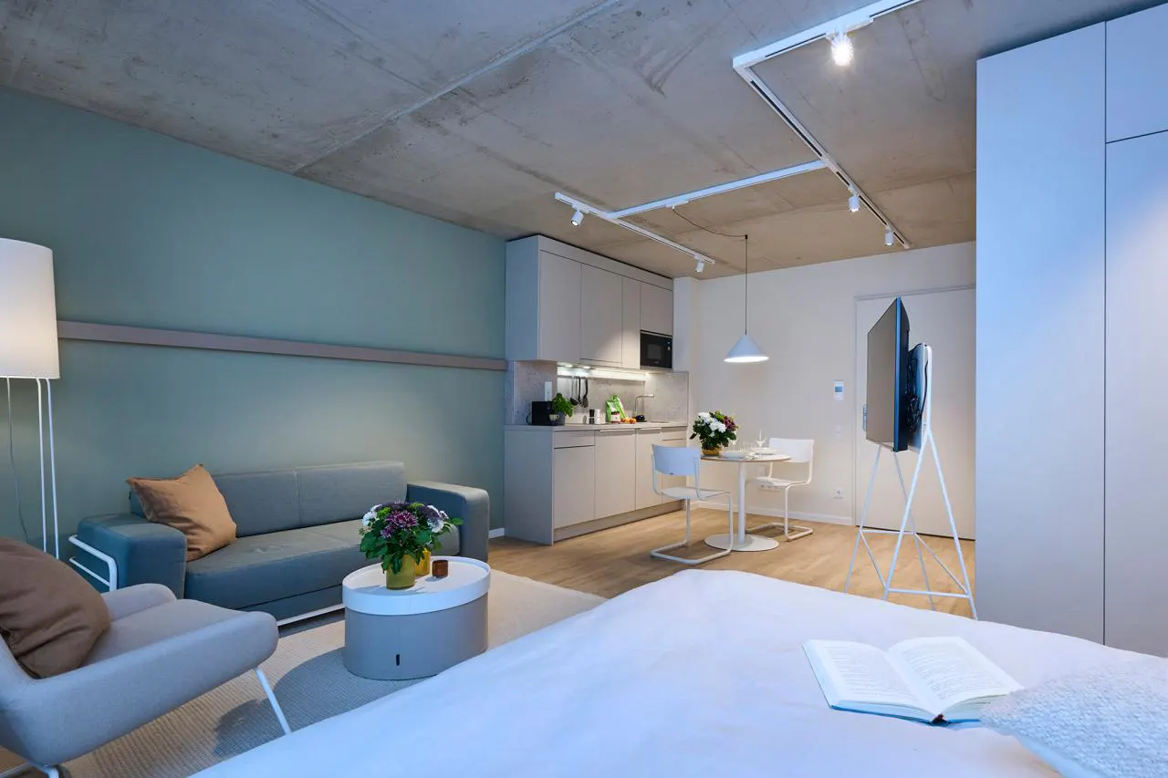 Studio apartment with a bed, sofa, chair, kitchen, and dining table. A book is open on the bed. The walls are green and the ceiling is concrete.