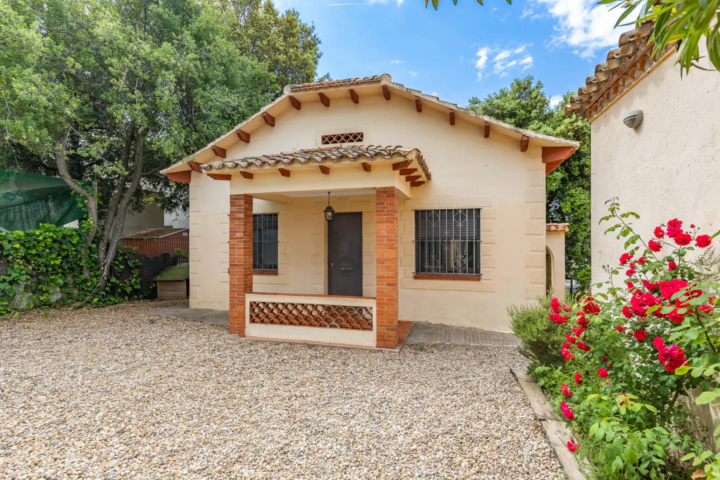 Beige stucco house with brick trim, a tile roof, and a gravel yard. Red roses bloom near a walkway.