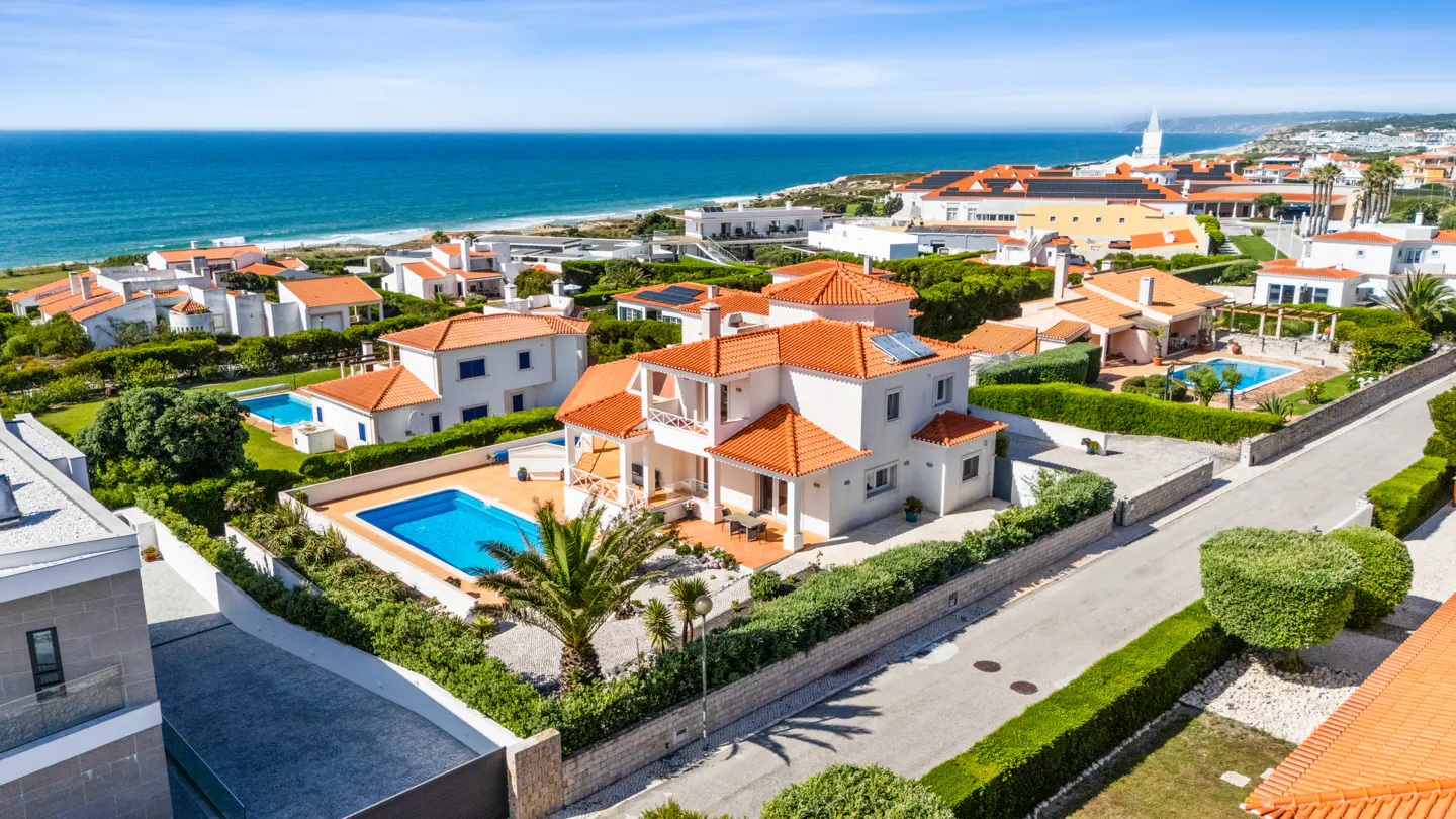 Aerial view of white houses with orange roofs, pools, and ocean backdrop under a blue sky.