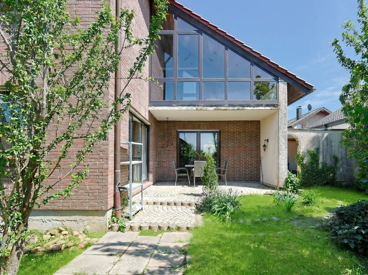 Brick house exterior with a patio, table, and chairs. A large window is above the patio. Green grass and trees surround the house.