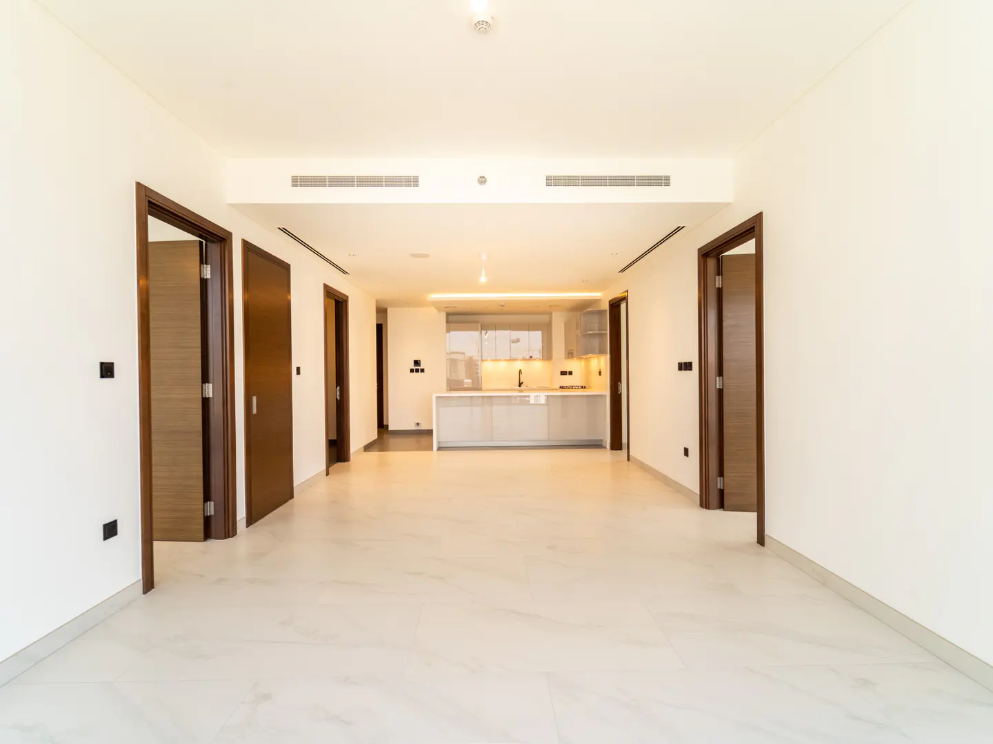 Modern open-plan living area with white marble floors, white walls, dark wood doors, and a distant kitchen with island.