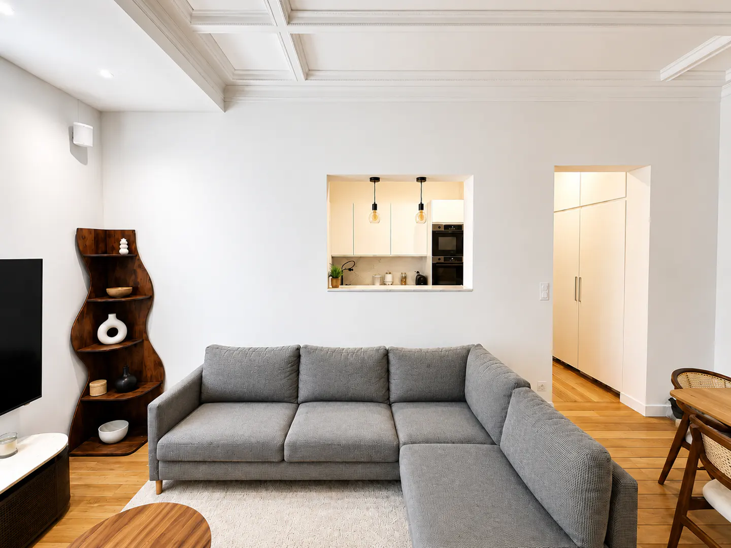 Bright living room with gray sectional sofa, wood floors, and white walls. A pass-through reveals a kitchen with white cabinets. A dark wood shelf adds visual interest.