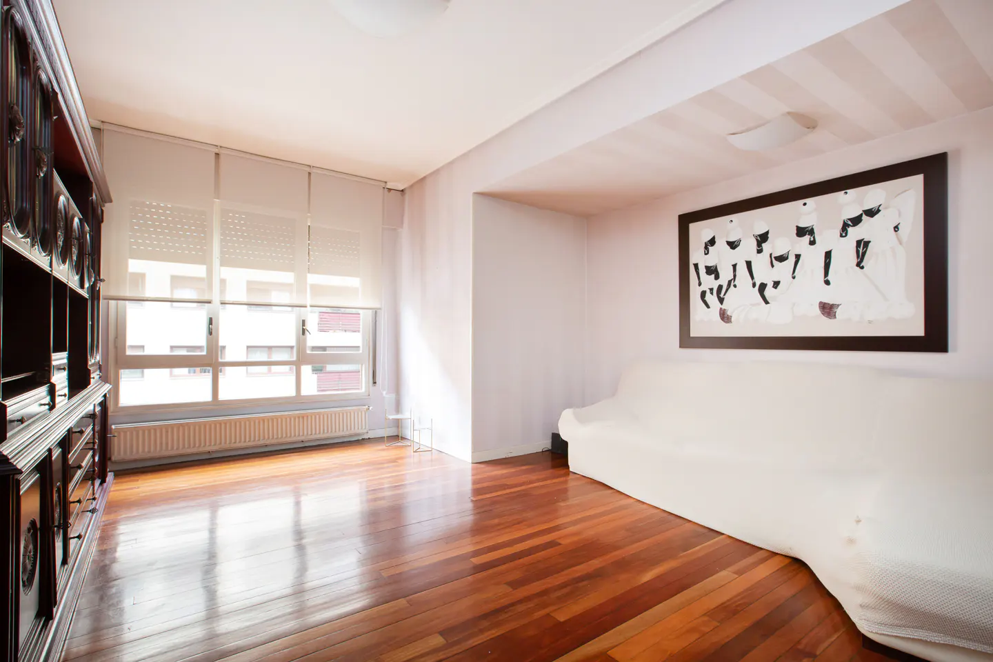 Bright living room with hardwood floors, white walls, and a large window with blinds. A dark wood cabinet stands against one wall, and a white sofa sits beneath framed art.