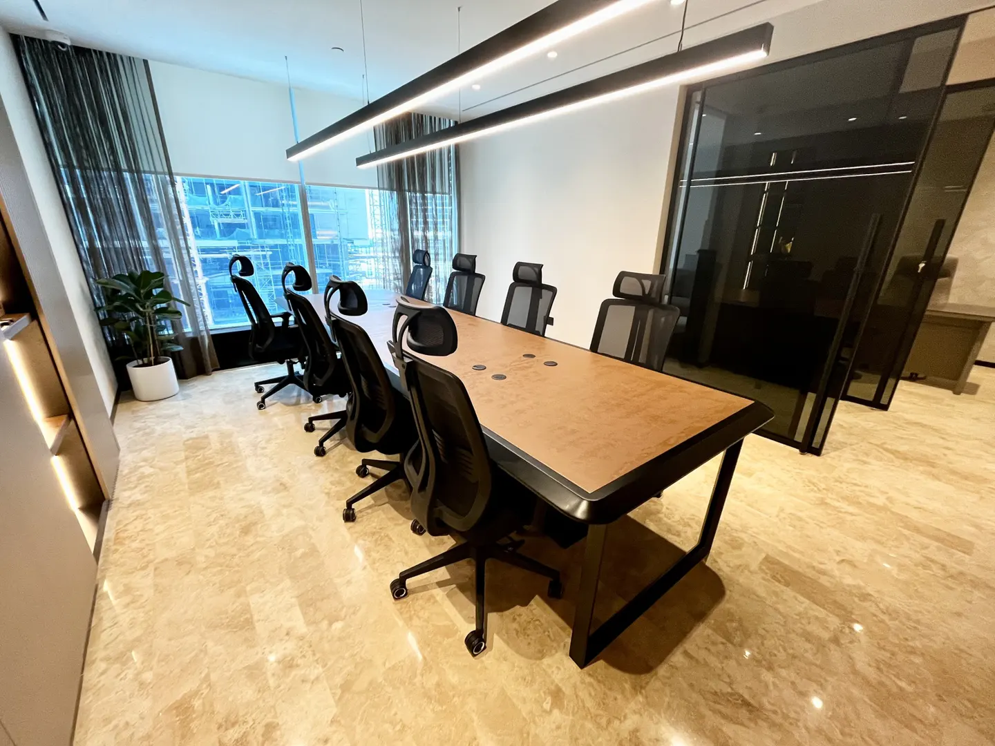 Conference room with a long wooden table, black chairs, and large windows with city views. Modern lighting fixtures hang above.