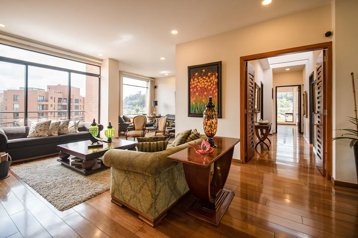 Bright living room with hardwood floors, a large window, and a brown sofa with decorative pillows. A painting hangs on the wall near an open doorway.