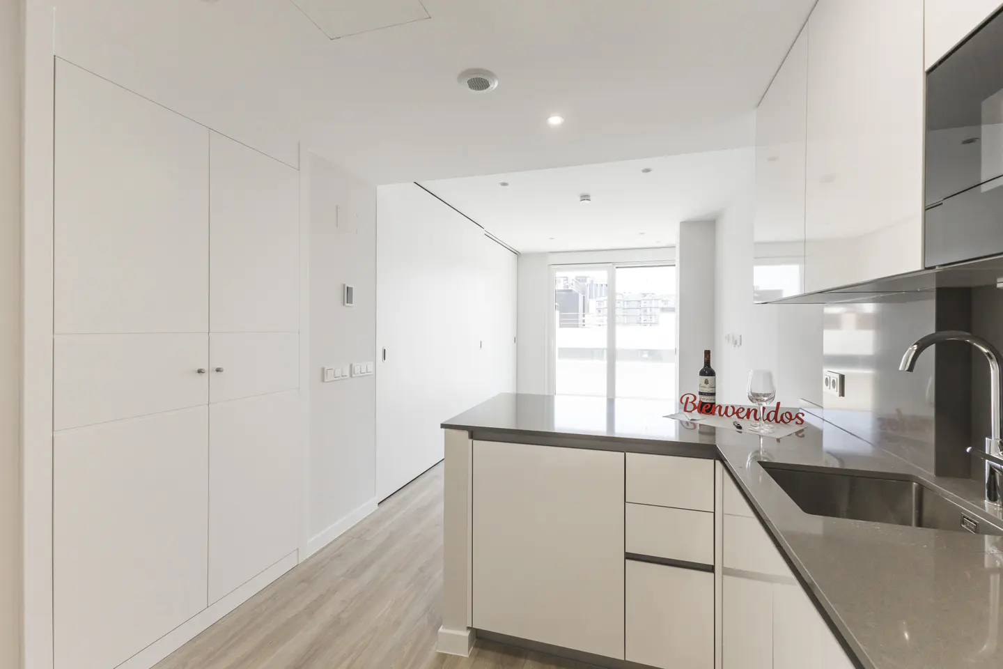Modern white kitchen with gray countertops, a wine bottle, and a "Bienvenidos" sign on the island, leading to a balcony.