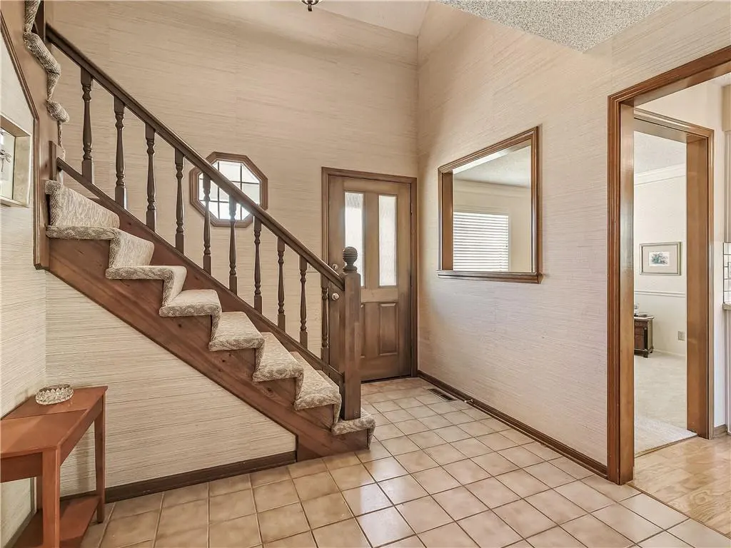 Foyer with wood staircase, beige carpet treads, and a small table. A wood door and a window are visible. The floor is tiled.