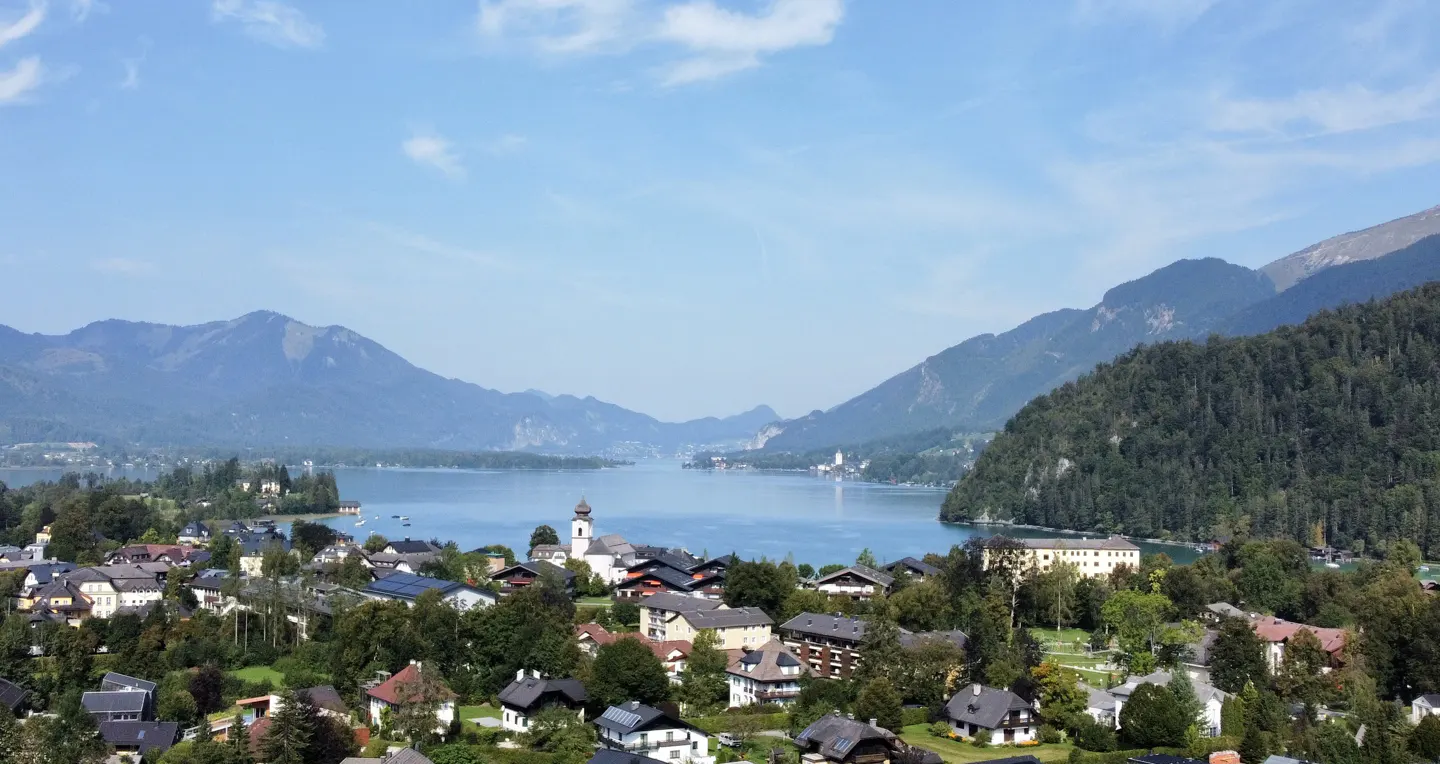 Scenic view of a village nestled by a lake, surrounded by mountains under a blue sky.