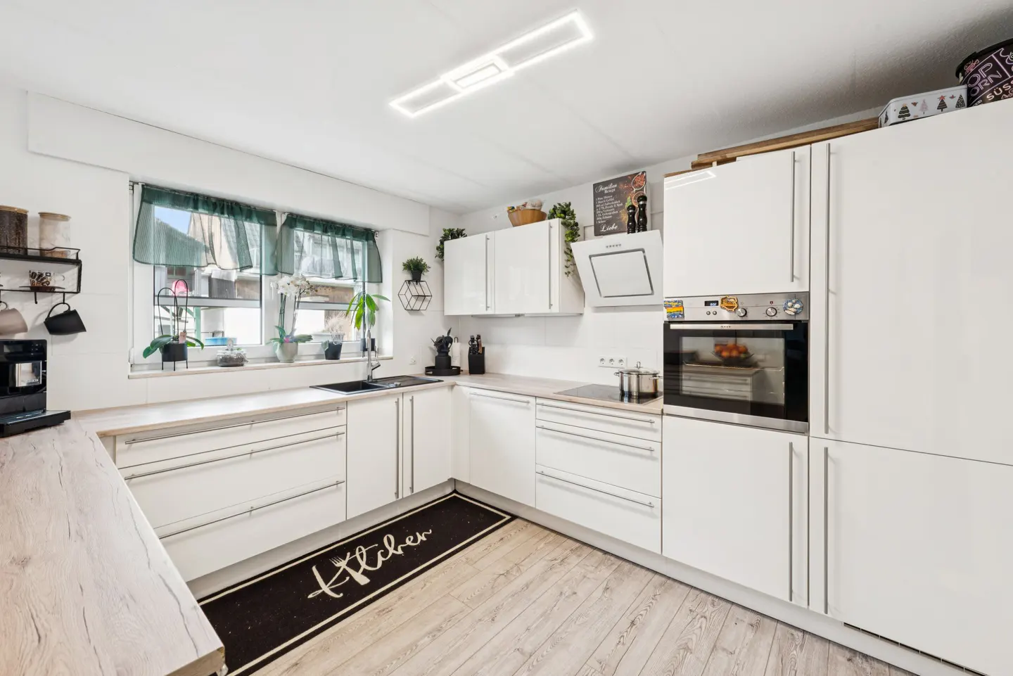 Bright kitchen with white cabinets, light wood floors, and a black "Kitchen" rug. A window with green curtains sits above the sink.