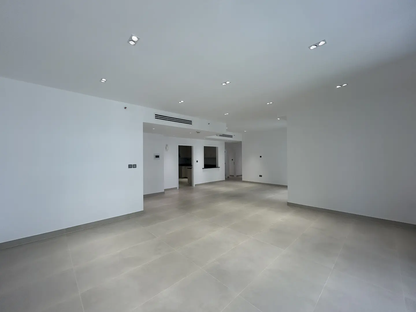 Spacious, empty living area with white walls, recessed ceiling lights, and light-colored tiled flooring.