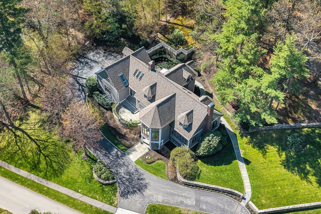 Aerial view of a gray house with a brown roof, surrounded by green trees and a lawn. A driveway leads to the house.