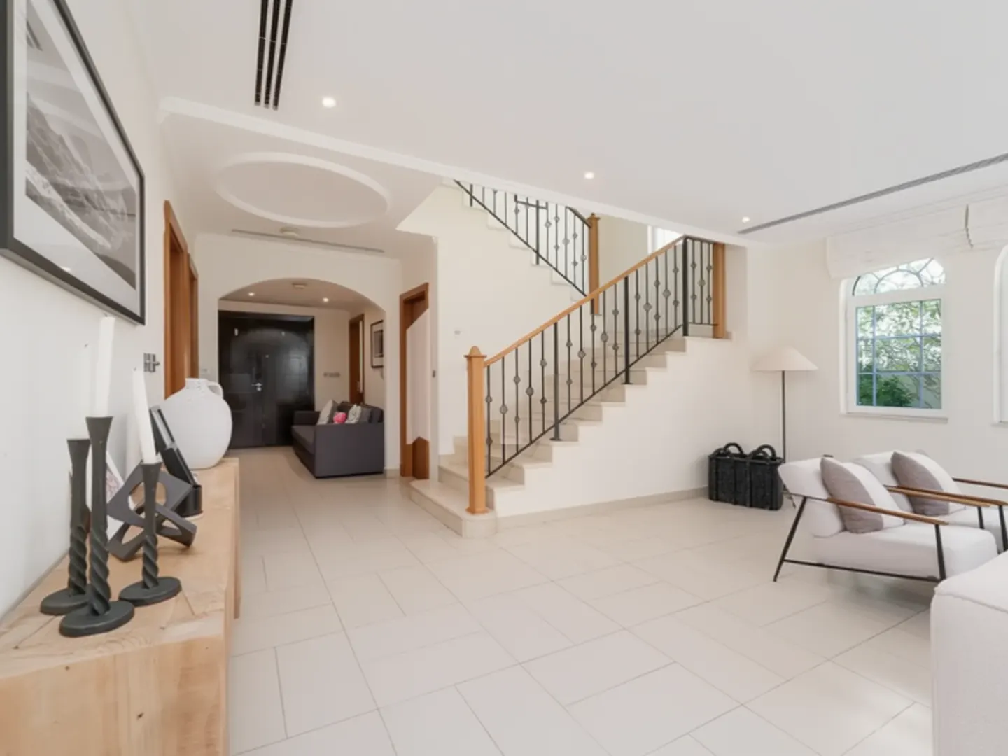Bright, open foyer with white tile floors, stairs with black iron railing, and seating area with white chairs near arched windows.
