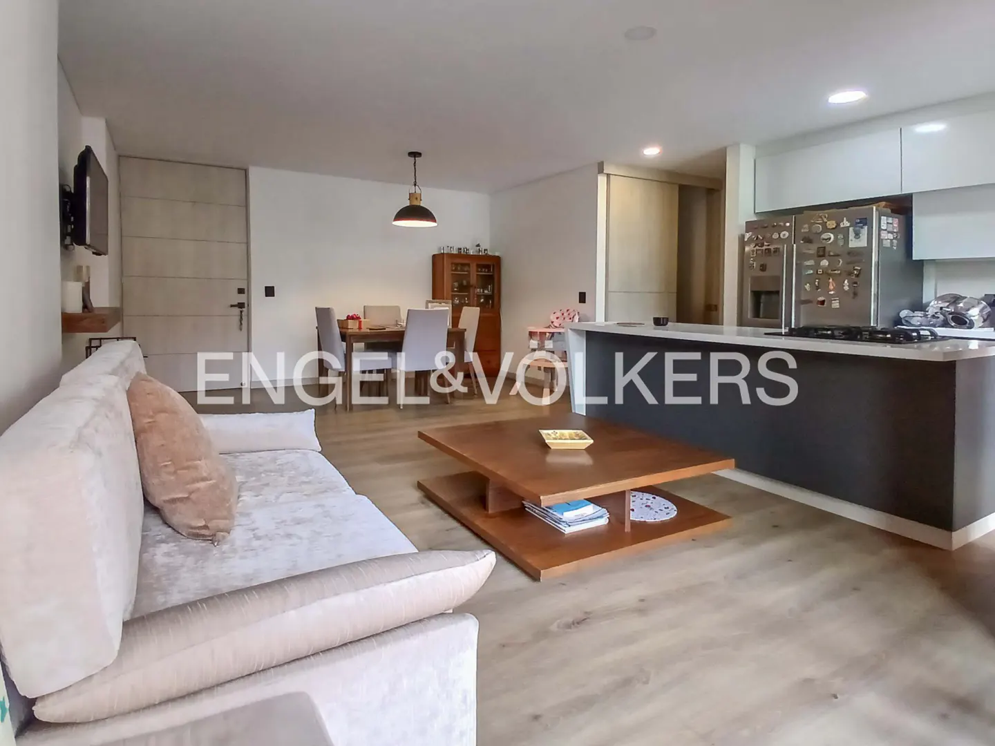 Open-concept living space with a sofa, wood coffee table, dining set, and kitchen island. Light wood floors and white walls.