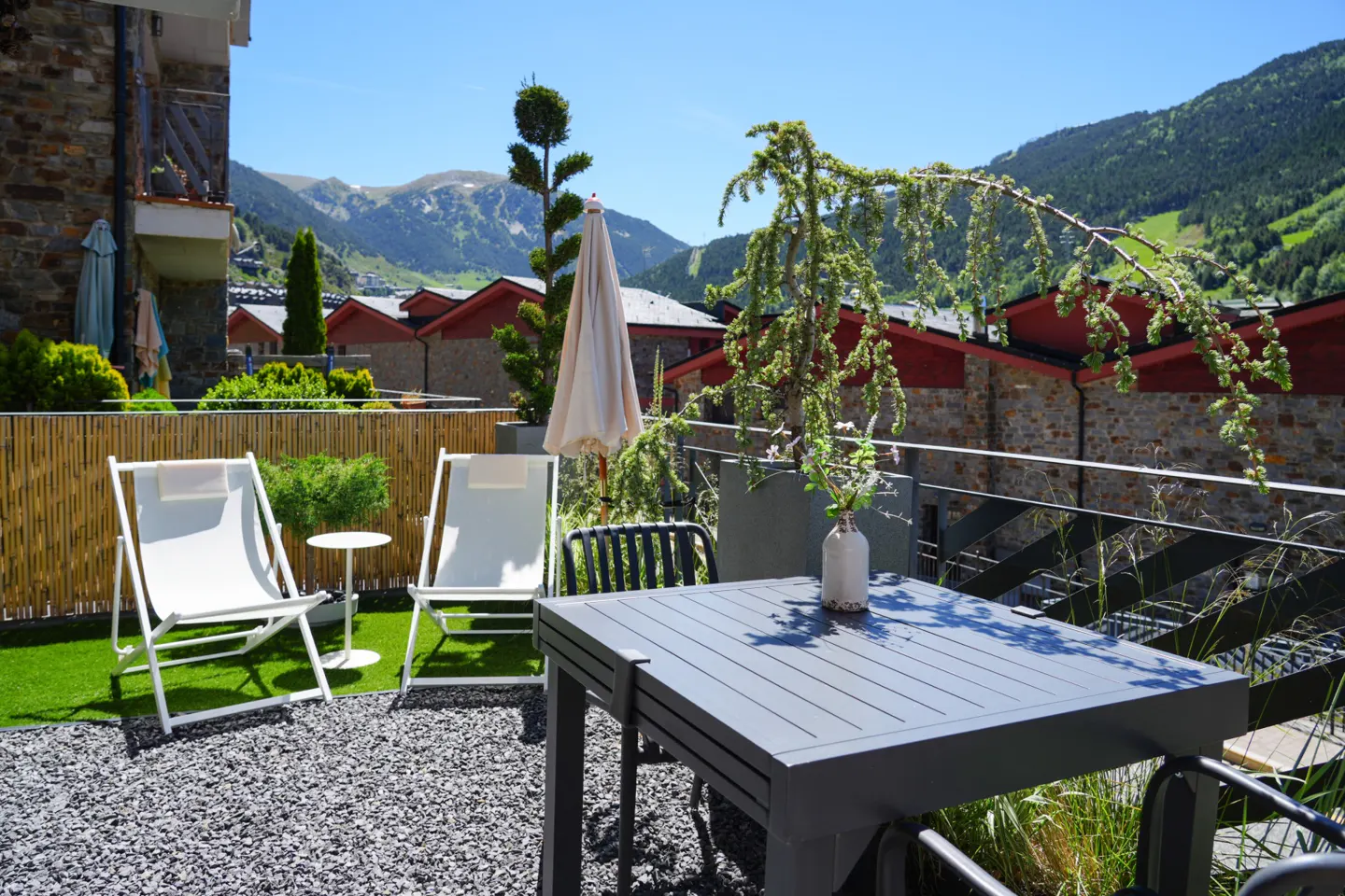 Sunny terrace with white lounge chairs, a dark table, and a small tree, overlooking a mountain village and green hills.