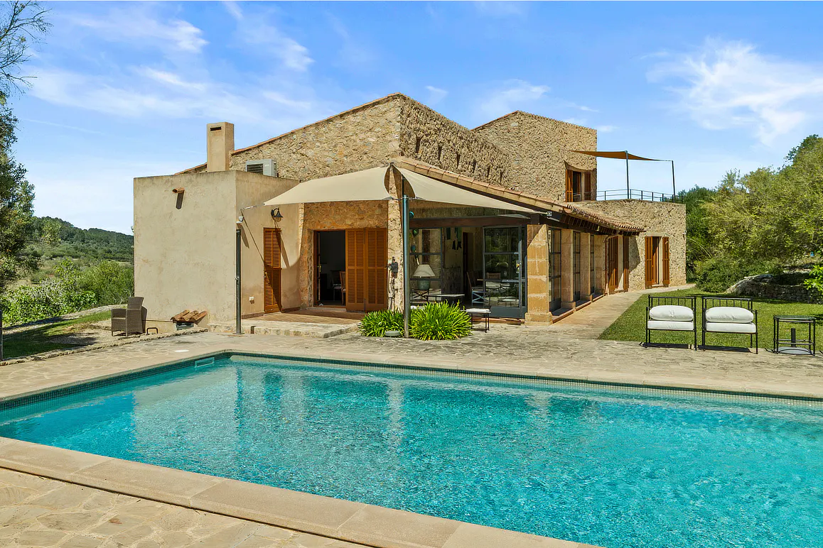 Exterior view of a stone house with a blue swimming pool in the foreground on a sunny day.