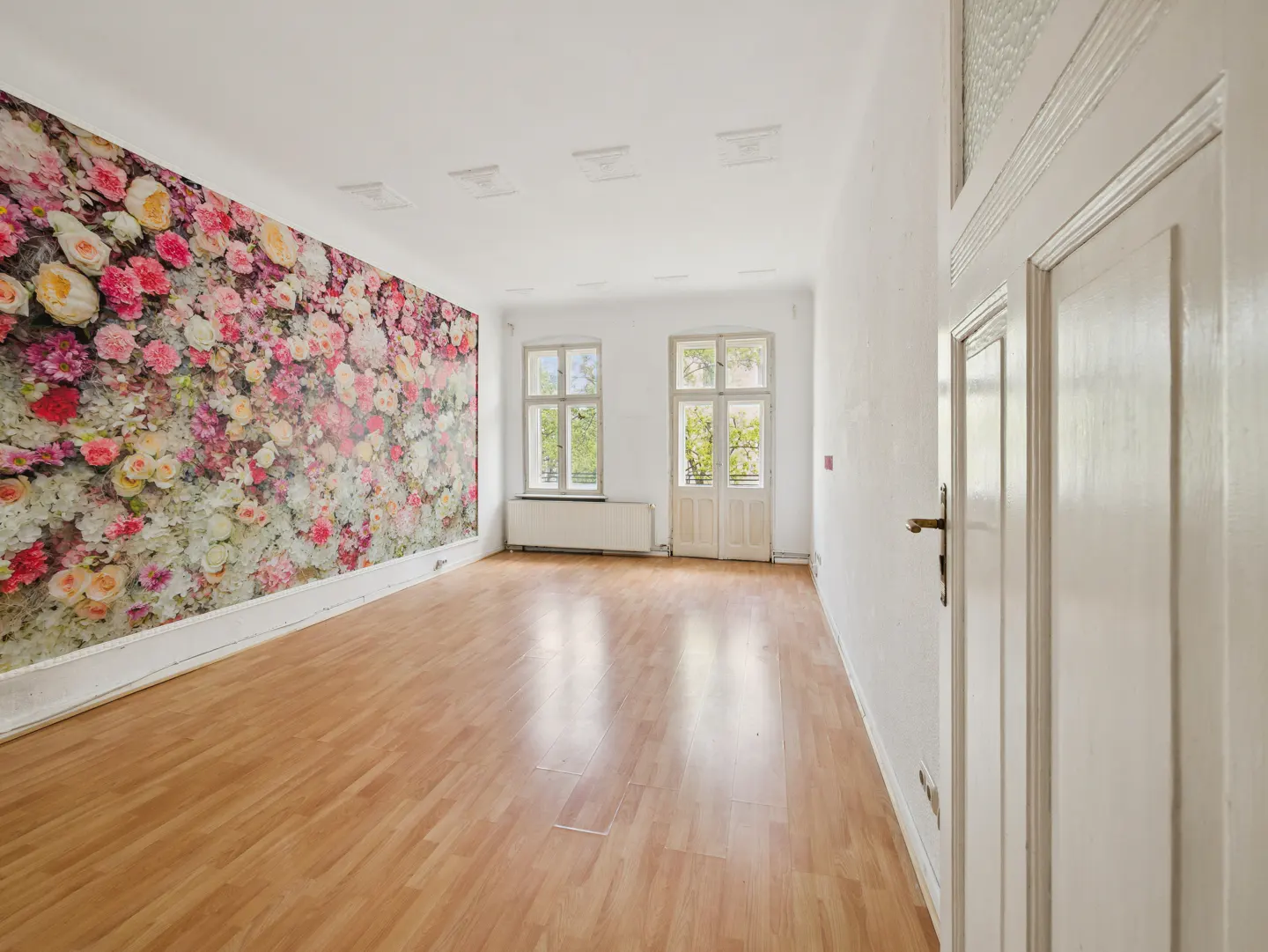 Bright, empty room with wood floors, white walls, and a floral wallpaper accent wall. Two windows let in natural light. A white door is partially open.
