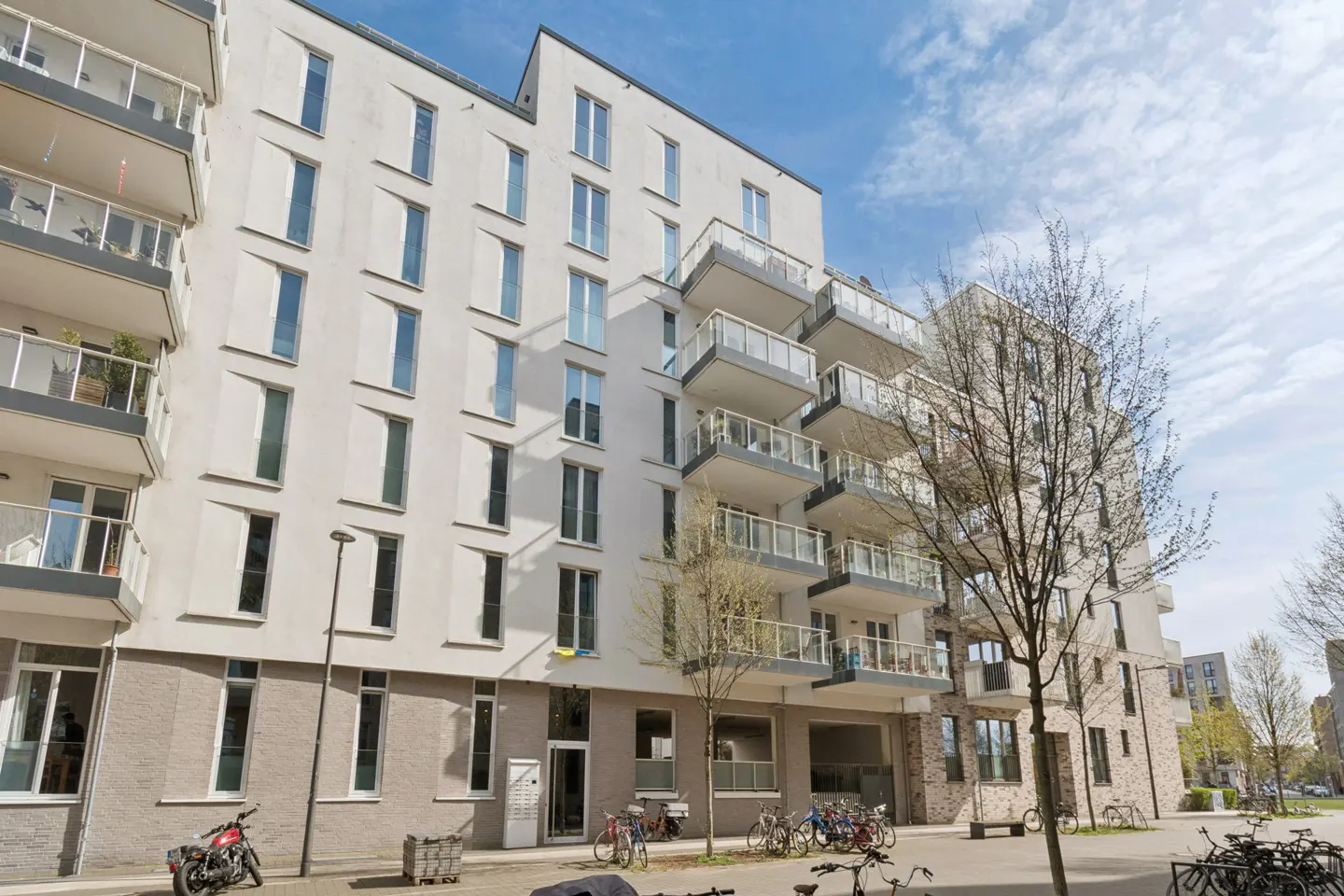 Modern apartment building with white facade, balconies, and many windows under a blue sky. Bicycles parked in front.