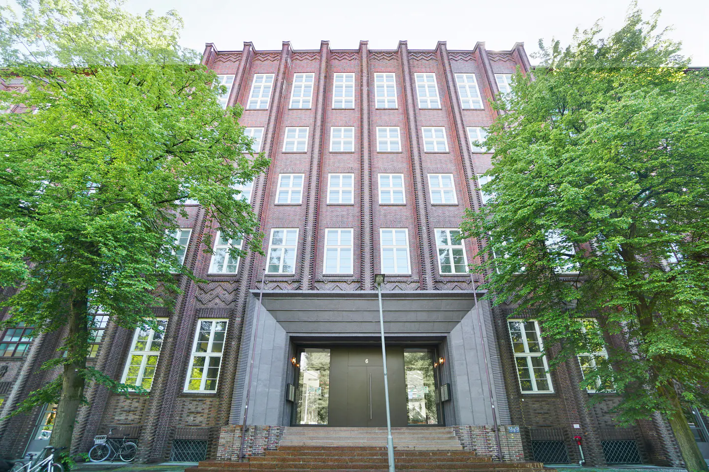 Exterior view of a tall brick building with many windows and a gray entrance framed by green trees.