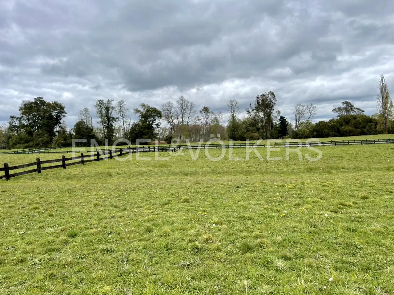 A green field with a wooden fence under a cloudy sky. Trees line the horizon.