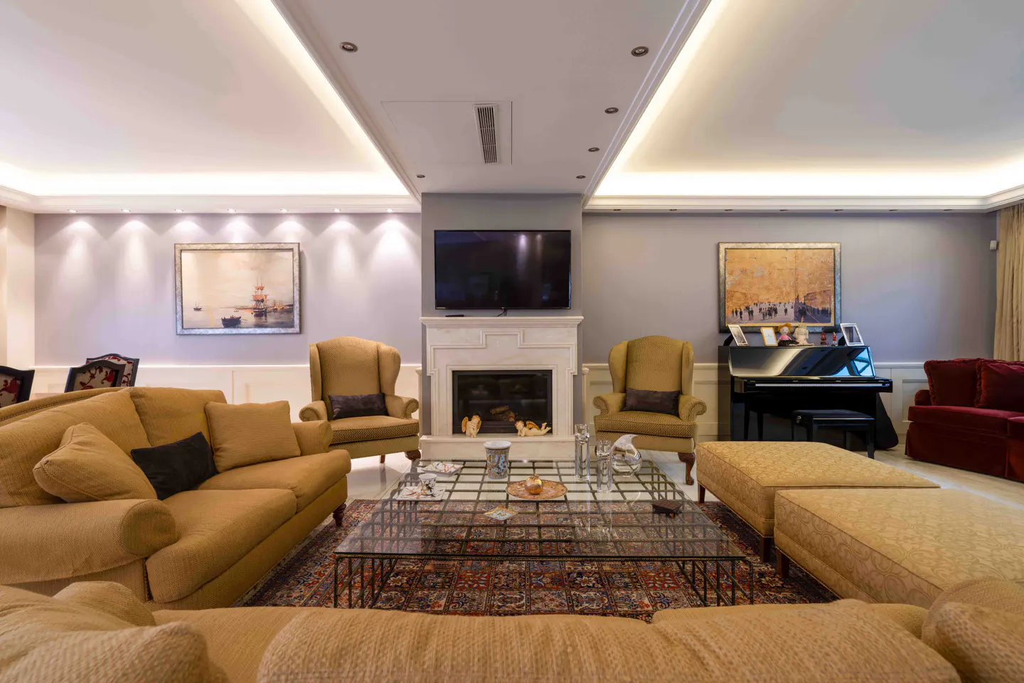 Living room with tan sofas, wingback chairs, and a glass coffee table on a patterned rug. A fireplace with a TV above it is flanked by artwork and a piano.