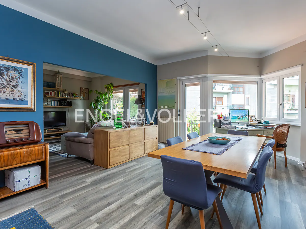 Open-concept living and dining area with blue accent wall, wood floors, and a large dining table with blue chairs.