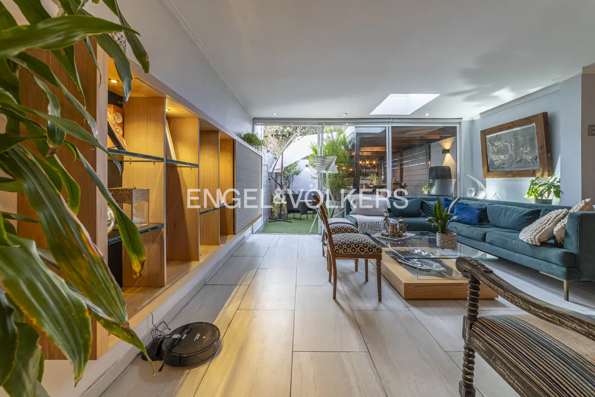 Bright living room with a blue sofa, glass table, and chairs. A bookshelf and a robot vacuum are on the left. A patio is visible through glass doors.