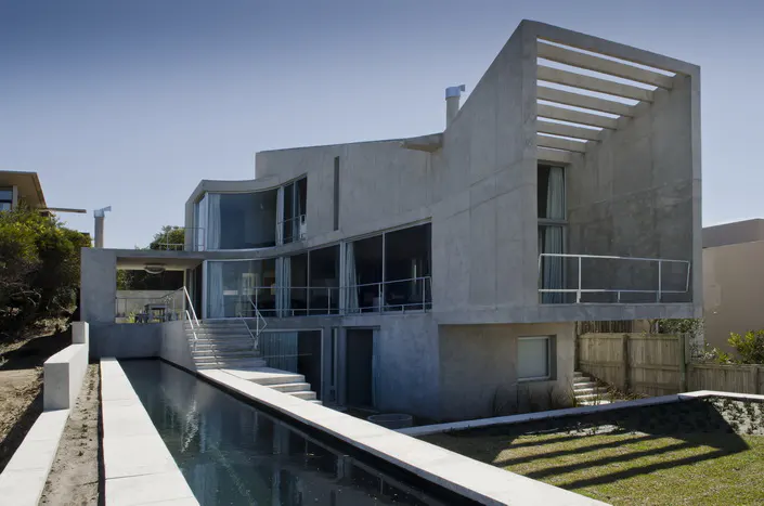 Modern gray concrete house with a long, narrow pool in front on a sunny day.